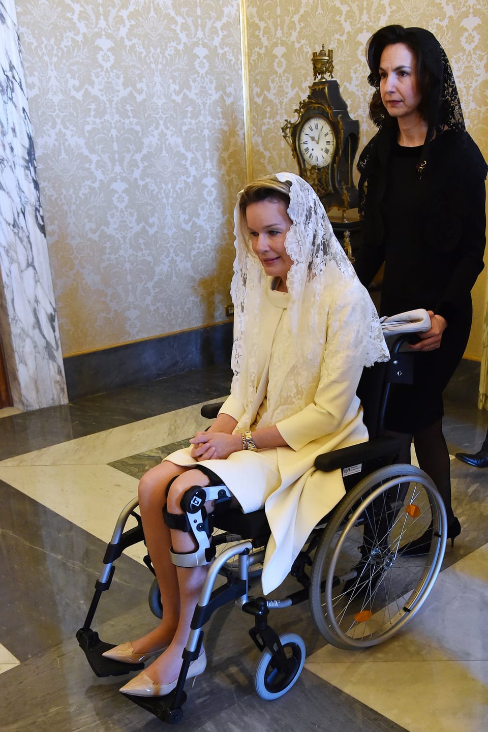 Königin Mathilde wird im Rollstuhl geschoben.