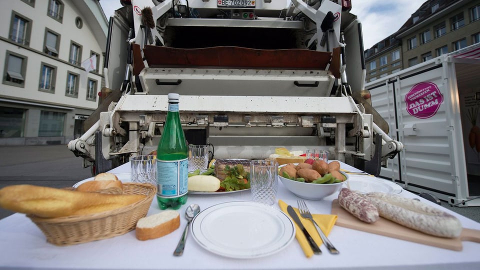 Zu sehen ist ein gedeckter Tisch mit Brot, Wurst, Kartoffeln, Käse, Salat und Wasser. Der Tisch steht in einer von Wohnhäusern gesäumten Strasse vor dem Schlund eines Müll-Wagens.