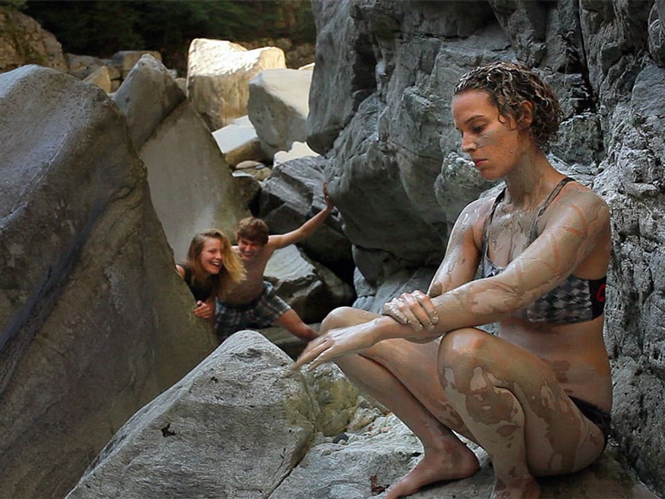 Eine junge Frau im Bikini sitzt auf einem streicht sich mit Erde ein, hinter ihr schaut ein junges Paar hinter einem Stein hervor und lacht.