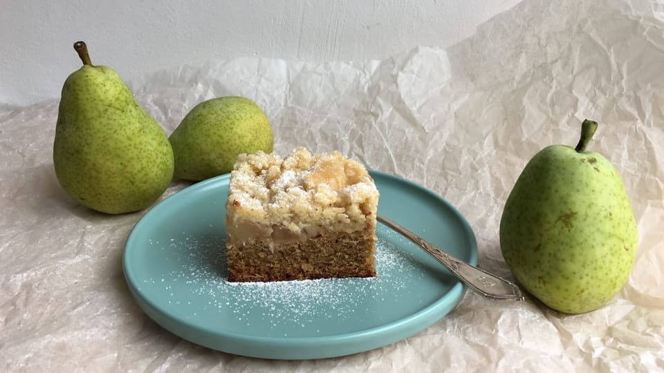 Ein rechteckiges Stück Birnenkuchen mit Crumbe auf einem graublauen Teller. Im Hintergrund drei grüne Birnen.