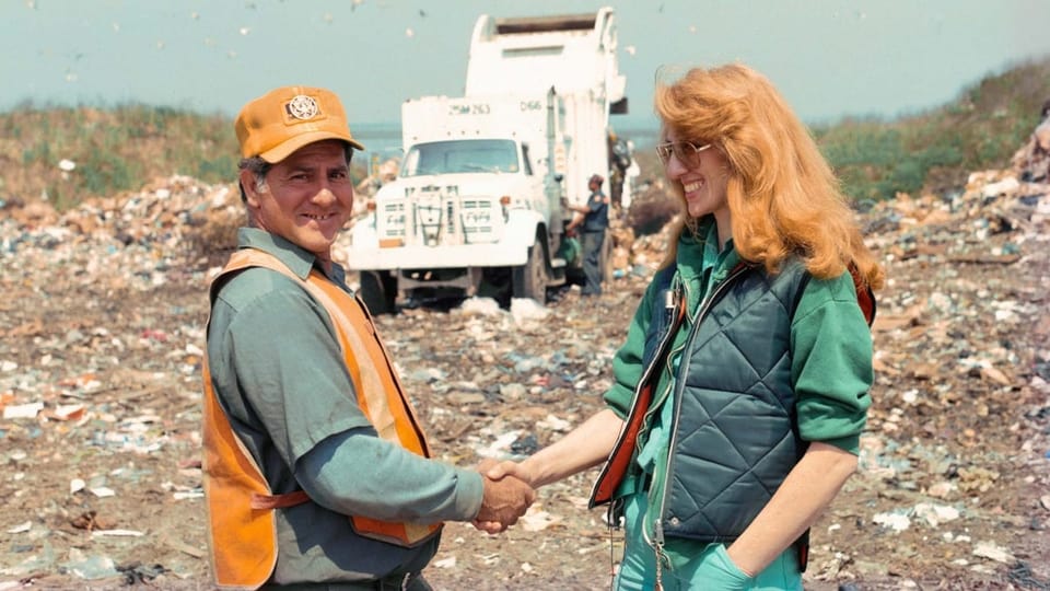 A woman shakes hands with a garbage disposal man, both laughing, in the background a garbage truck.