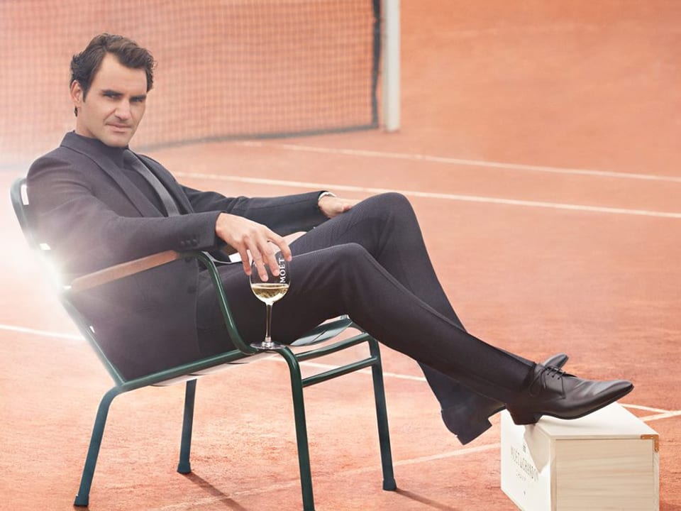 Roger Federer sitzt im Anzug auf einem Stuhl auf dem Tennisplatz, hält ein Champagner-Glas in der Hand und hält die Füsse auf eine Holzkiste.