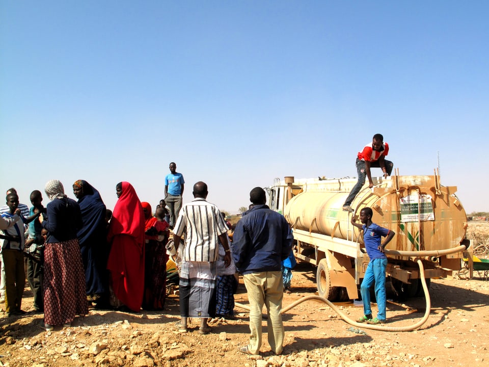 Um einen Tankwagen in der Wüste stehen Frauen in farbigen Gewändern und Männer, die einen Wasserschlauch auslegen.