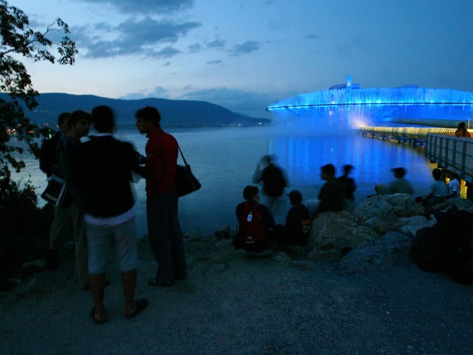 Menschen am Seeufer blicken auf eine beleuchtete Installation.
