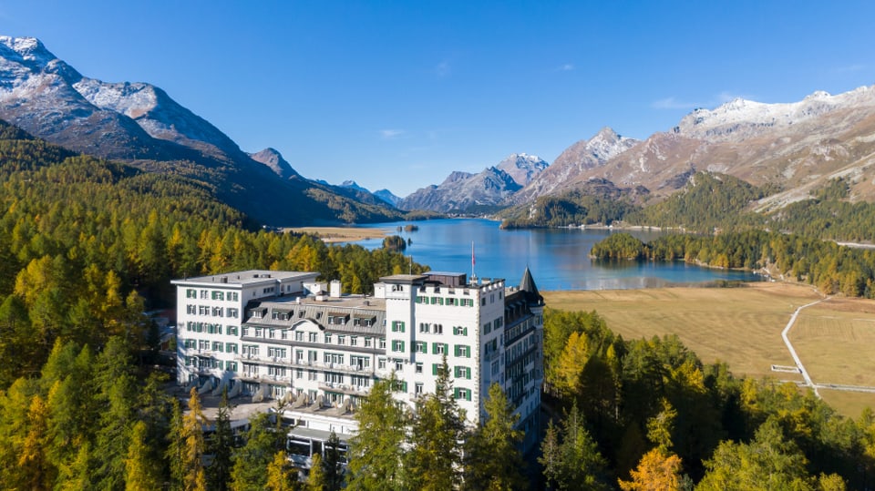 Ein Grand Hotel thront im Wald über einem See. Im Hintergrund berge und blauer Himmel.