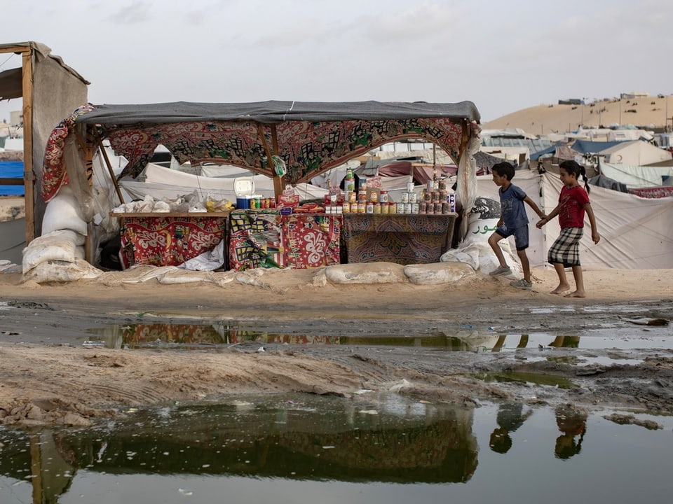 Kinder laufen an einem Marktstand vorbei in einem Flüchtlingslager mit Reflexion im Wasser.