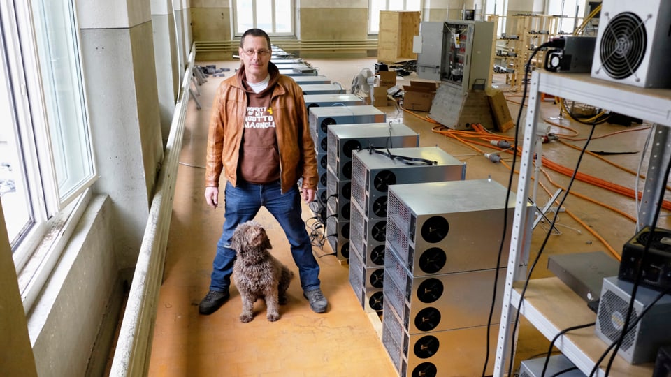 Ein Mann und sein Hund in einer Fabrikhalle; rechts Racks mit Rechnern.