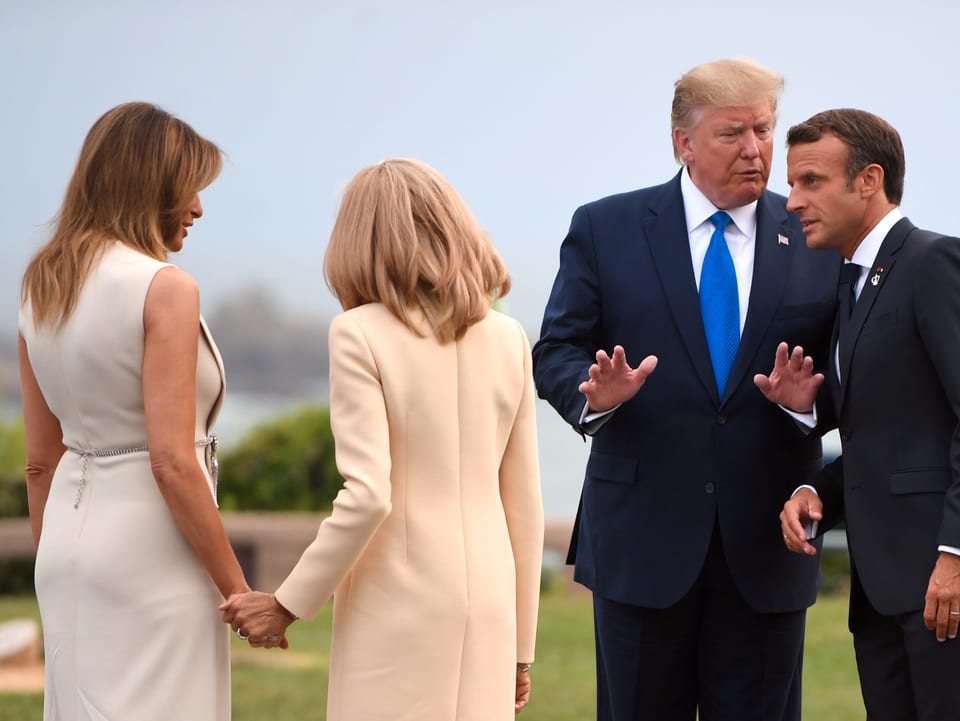 Melanie Trump und Frau von Macron geben sich die Hand und tragen beide ein cremfarbiges Kleid. Daneben unterhalten sich Trump und Macron.