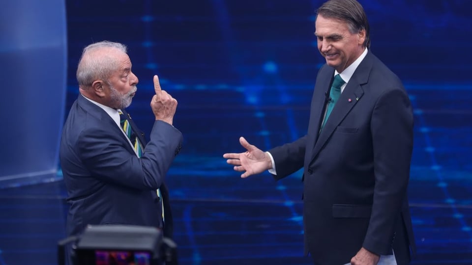 Lula hebt den Finger, während Bolsonaro daneben nach unten schaut, lacht und dabei die Hand verständnislos etwas anhebt.