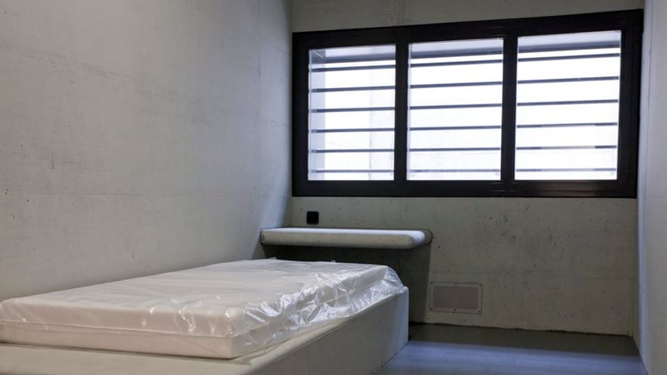 Eine Zelle mit Bett und vergittertem Fenster.