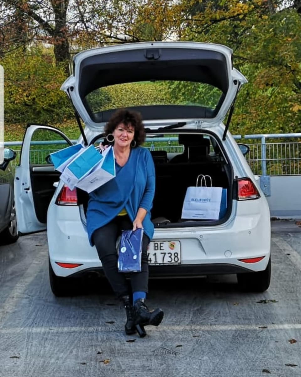 Eine Dame in einem blauen Cardigan sitzt beim offenen Kofferraum ihres weissen Autos, sie hält einige Papiertragetaschen in ihrer rechten Hand, über ihre Schulter geschwungen