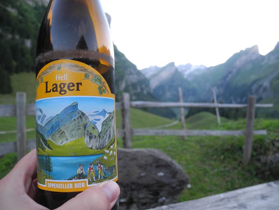 Bierflasche mit Sujet  wird vor Bergpanorama gehalten.