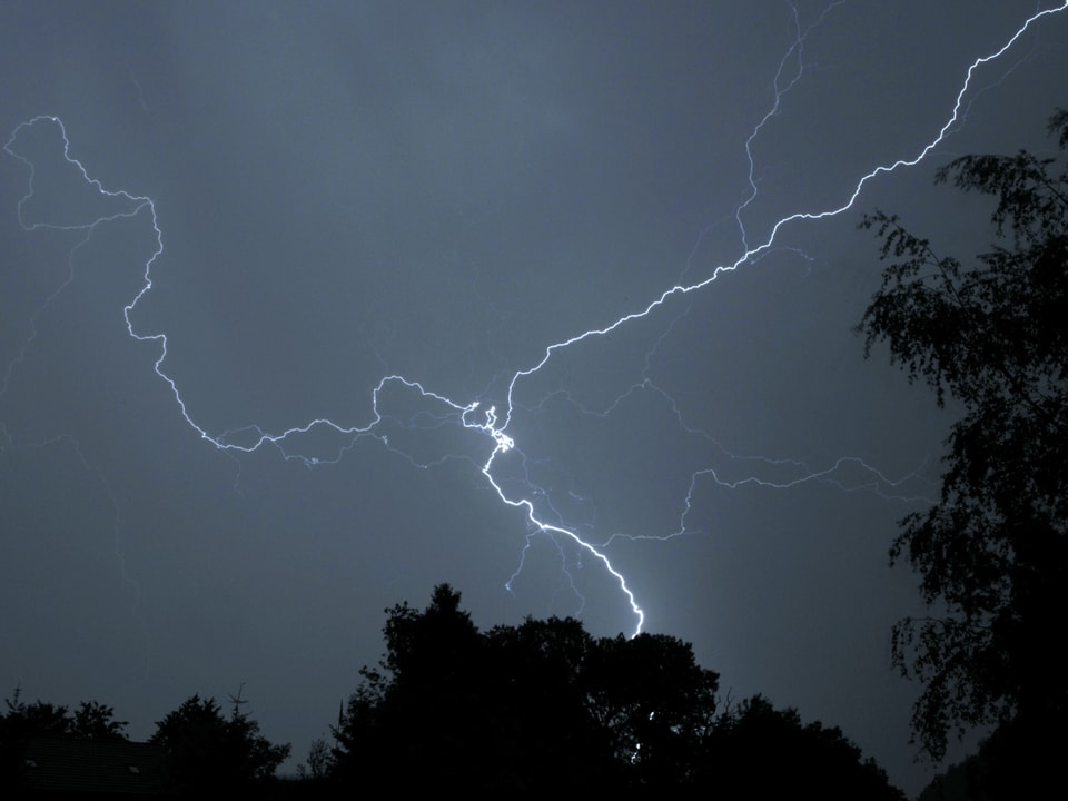 4. Juli Dunkler Himmel mit den feinen und hellen Linien eines Blitzes. Unten sind noch die Konturen von Bäumen zu sehen. Es ist dunkle Nacht.