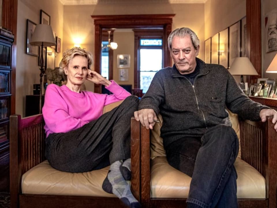 Älteres Eherpaar auf zwei Sesseln in Wohnzimmer. Frau mit pink farbigen Pullover, Mann in grauer Weste. 