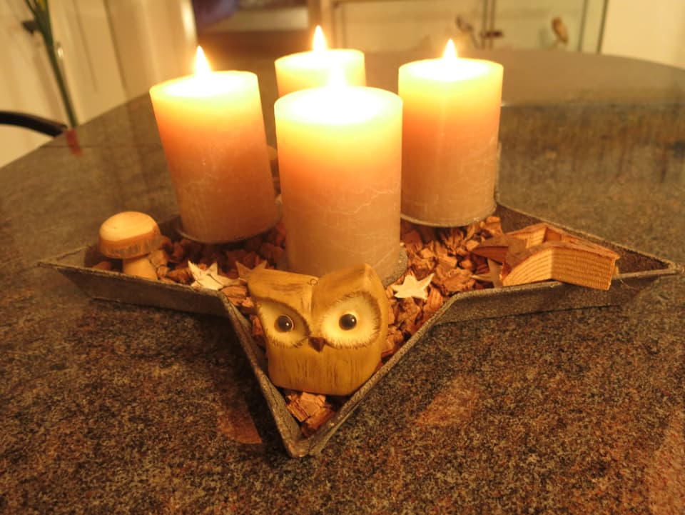 Ein Stern gefüllt mit Holzschnitzeln. Darauf stehen vier weisse Kerzen. Verziert ist das Ganze mit Holzfiguren.