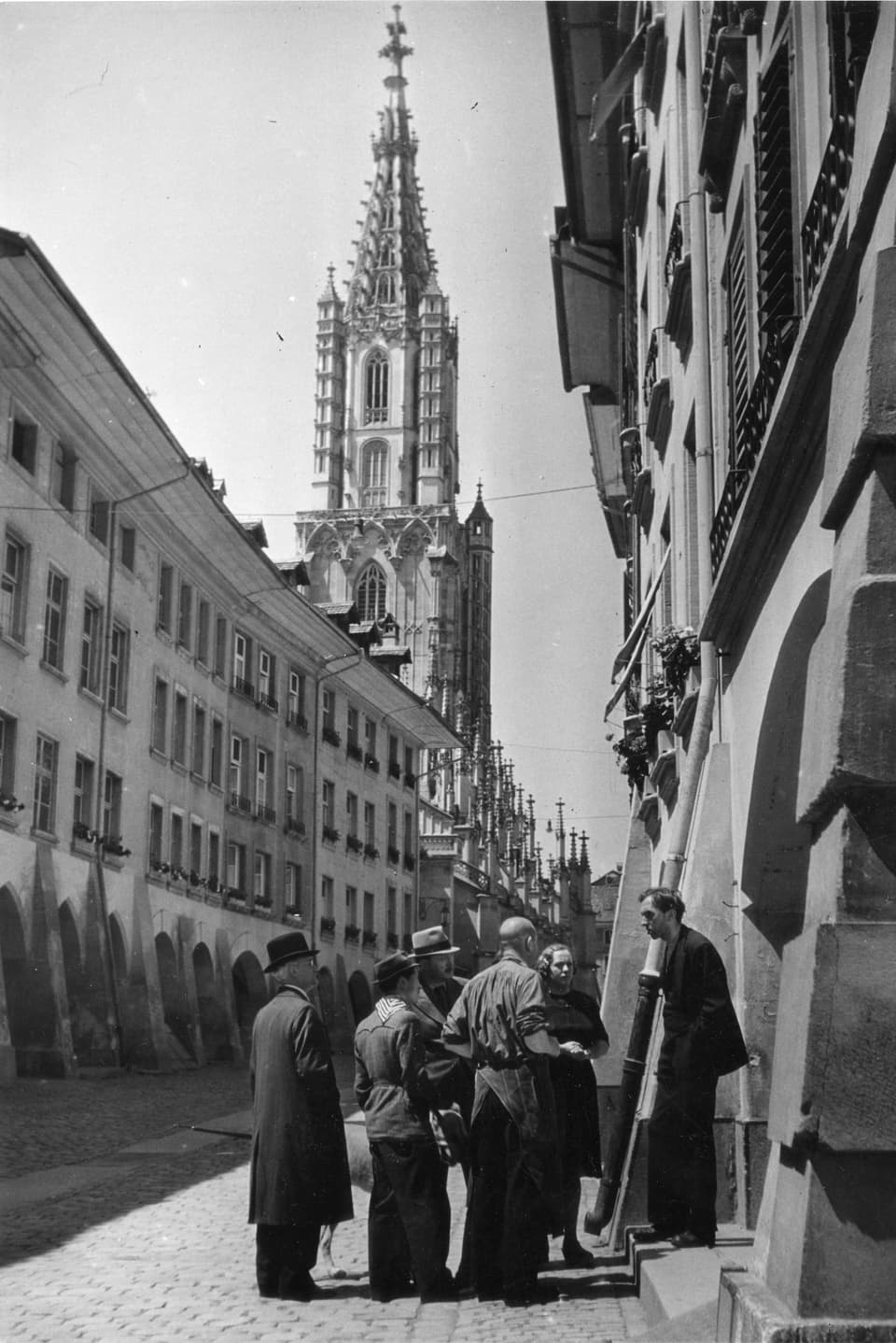 Vier Männer und zwei Frauen stehen an einem Haus in einer Strasse in Bern. Im Hintergrund steht das Berner Münster.