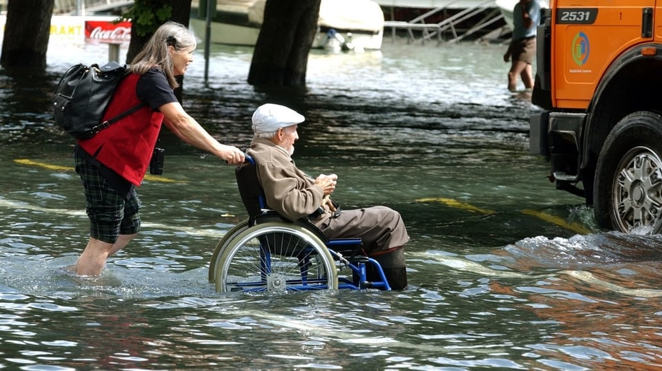Eine  Frau schiebt einen betagten Mann im Rollstuhl durch die überschwemmten Strassen