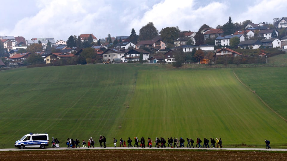 Flüchtlinge laufen hinter Polizeiwagen her, dahinter ein bayrisches Dorf. 