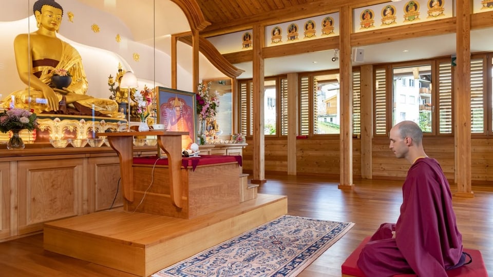 ein Herr mit Glatze sitzt in Mönchsrobe am Boden vor einer goldenen Buddhastatue