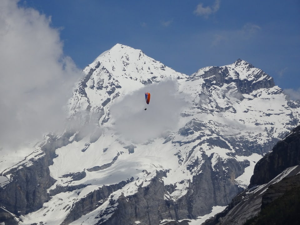 Ein Gleitschirmflieger vor dem verschneiten Blümlisalphorn. Kleine Quellwolken umgeben das Horn.
