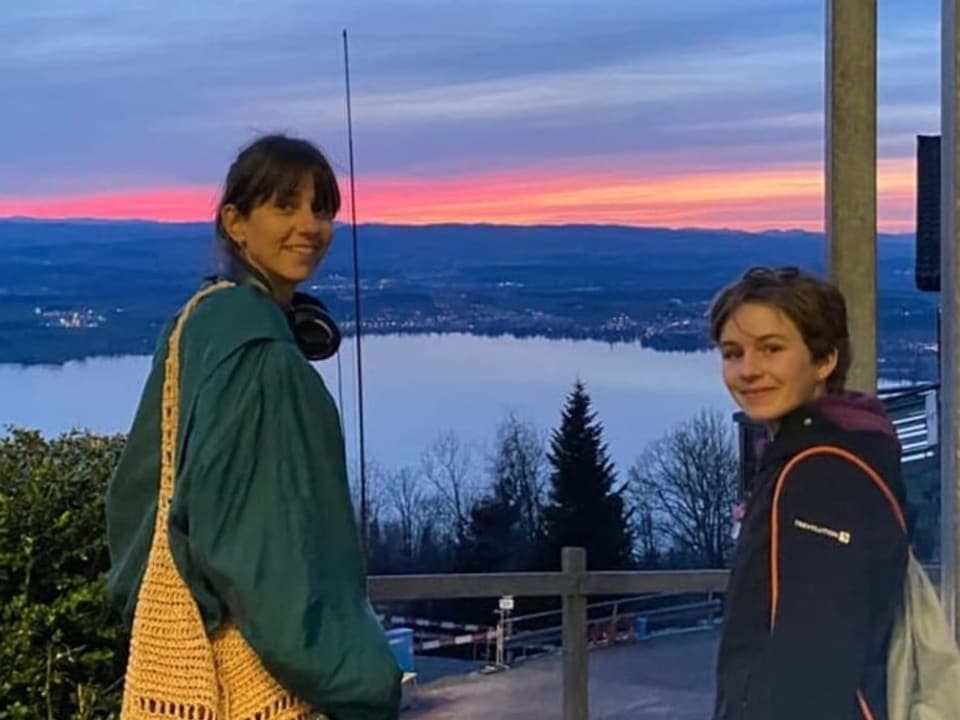 Links eine Frau in grüner Jacke und rechts ein Mädchen mit kurzen Haaren. Im Hintergrund sieht man Berge und einen See.