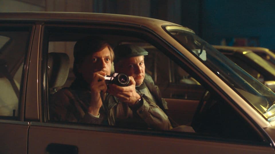Zwei Männer in einem Auto. Der eine von ihnen hält eine Kamera in der Hand. 
