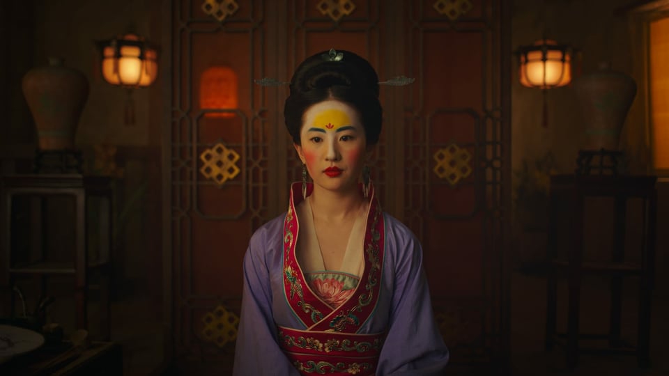 Mulan sitzt geschminkt und traditionell gekleidet an einem Tisch.