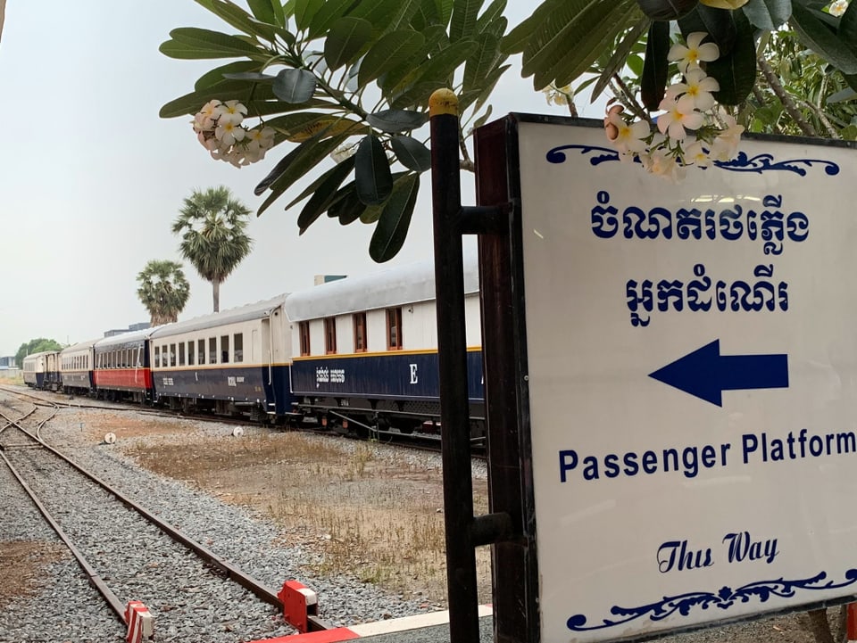 Im Vordergrund ein Informationsschild und Wegweiser. Im Hintergrund Palmenartige Pflanzen und ein wartender Zug der Royal Cambodian Railway.