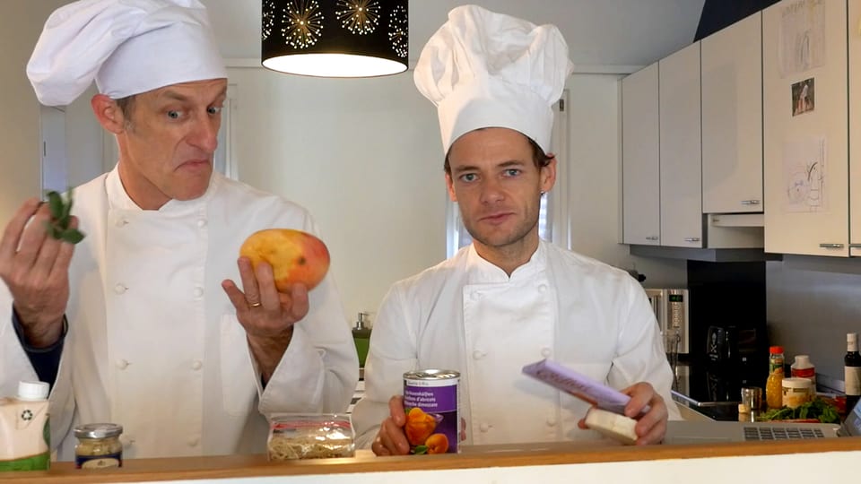 Zwei Männer in Kochkleidung halten Zutaten in die Kamera.