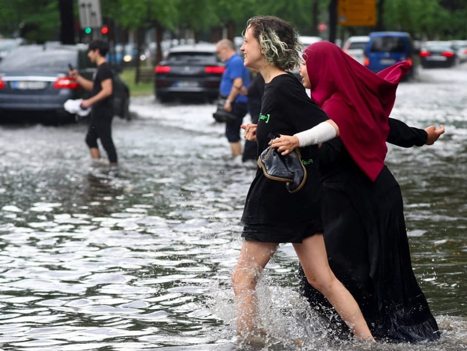 Zwei Menschen gehen auf einer Strasse, die knöchelhoch überflutet sind.