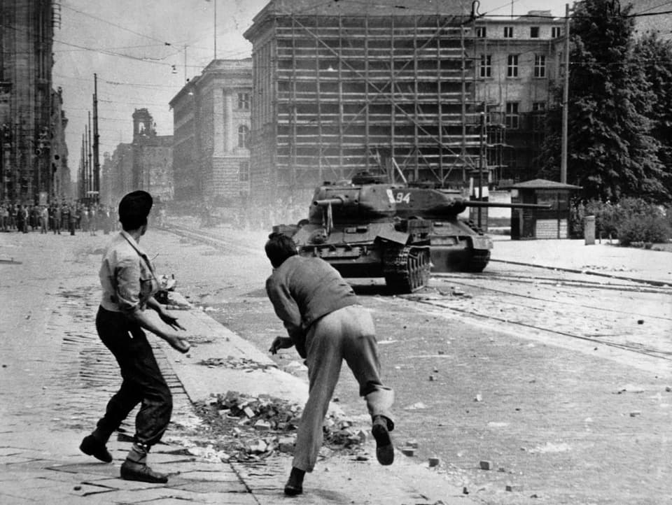 Schwarzweiss-Bild: Zwei Männer werfen Steine auf einen Panzer.