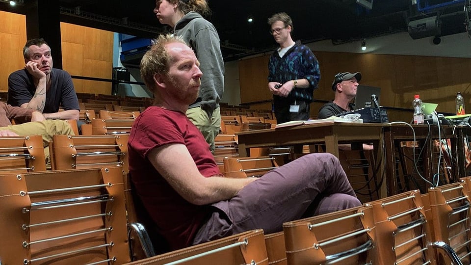 Eher junger Mann steht mit dunkelroter Mütze draussen vor Beton-Treppenhaus, rötlicher Dreitagebart