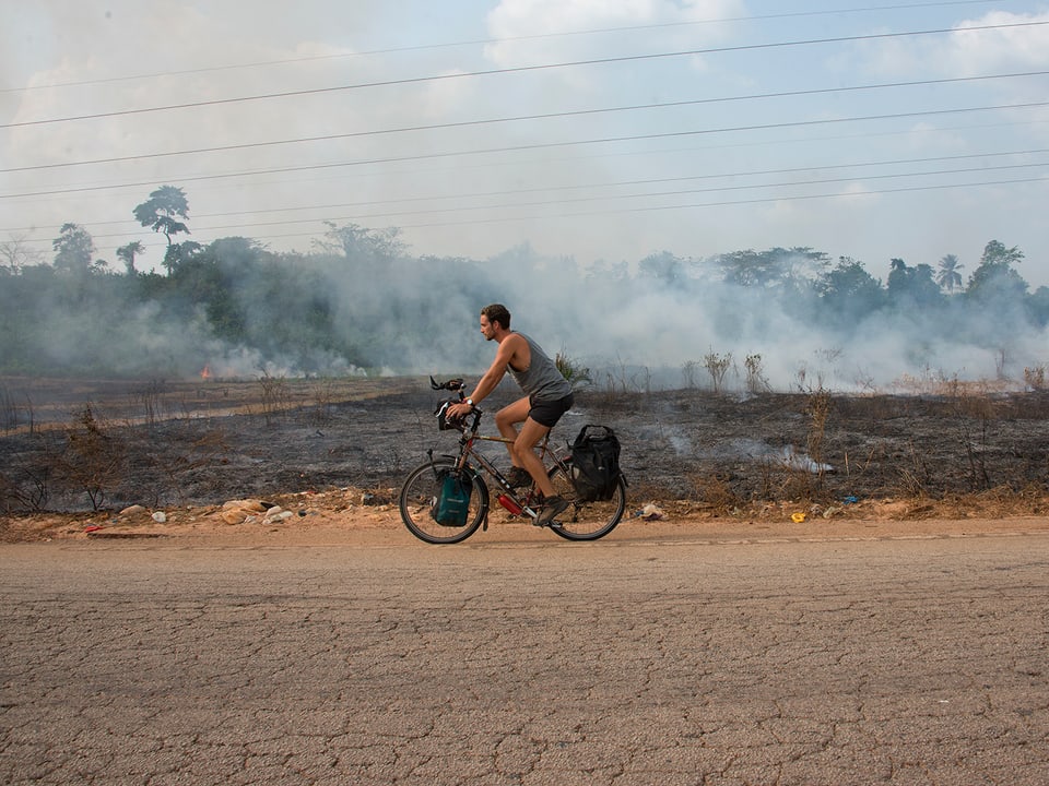 Lukas Biry fährt auf einer Strasse, im Hintergrund brennen und qualmen Büsche.