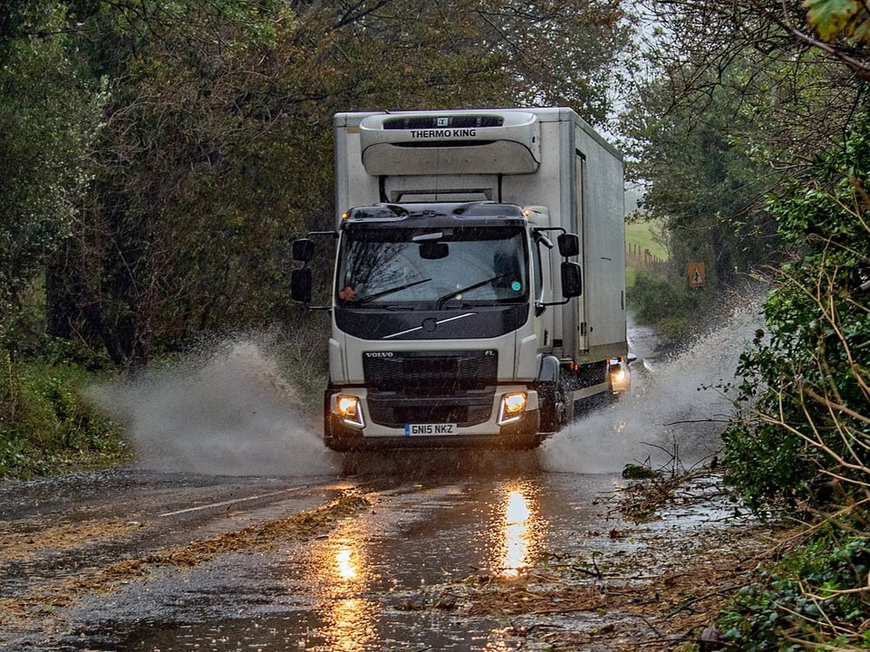 A truck is driving on a flooded road.  There are fallen branches all over the sides.