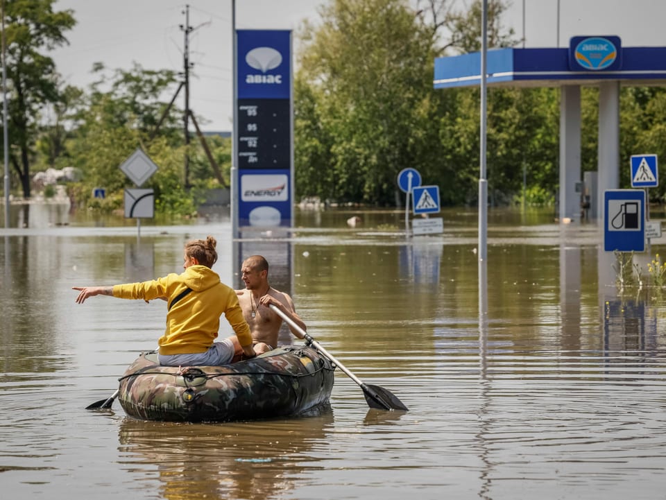Personen sitzen in einem Boot auf einer überschwemmten Strasse. Im Hintergrund steht eine Tankstelle unter Wasser.