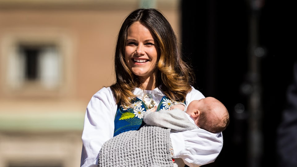 Prinzession Sofia hält in traditioneller Tracht (gelber Rock, weisse Bluse und geblümte Weste) ihren Sohn in den Händen, während ihr Ehemann der Menschenmenge zuwinkt.