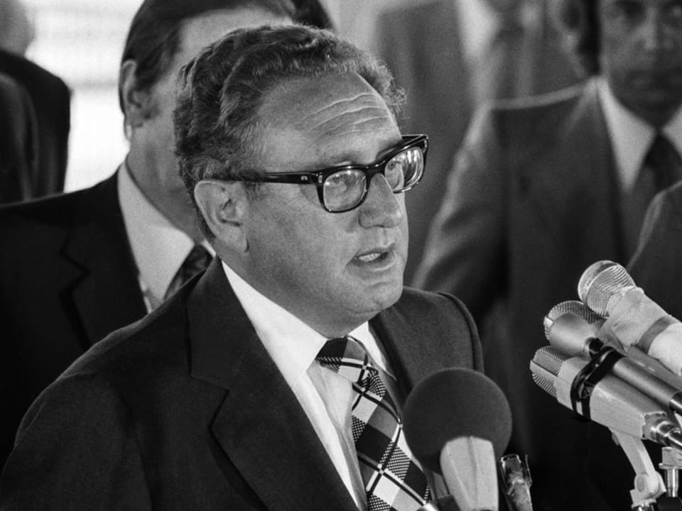Black and white photo of Henry Kissinger.  He speaks into different microphones.
