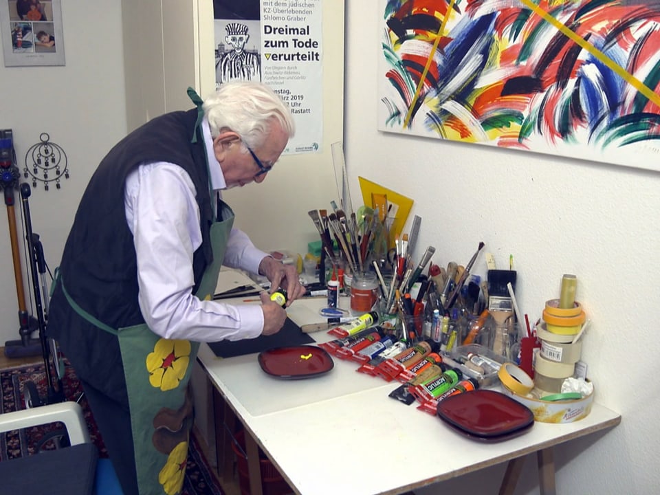alter Mann beugt sich über Tisch mit Farbe in der Hand, dahinter hängt ein Kunstwerk.