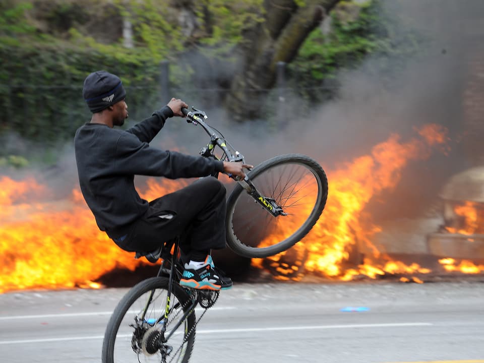 Ein Fahrradfahrer vor brennenden Polizeiwagen. 