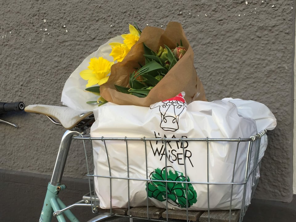 Ein Velokorb, angelehnt an eine beige Hausmauer, prall gefüllt mit einer Tasche voller Frischprodukte und zwei Blumensträussen.