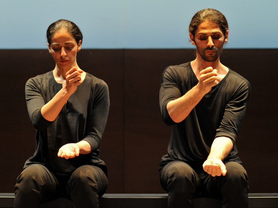 Eine Frau und ein Mann in schwarzer Alltagskleidung knien auf einer Bühne, und halten ihre Hände so, als würden sie töpfern.