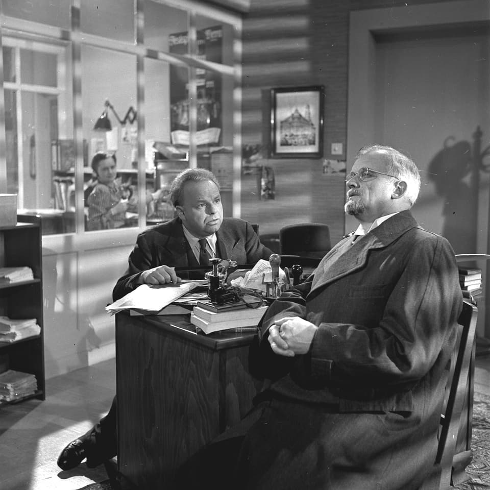 Ein Mann (Notar Tyffel) sitzt bei Redaktor Oppinger am Schreibtisch. Zweite Mann (Redaktor Oppinger) schaut Tyffel fragend an.