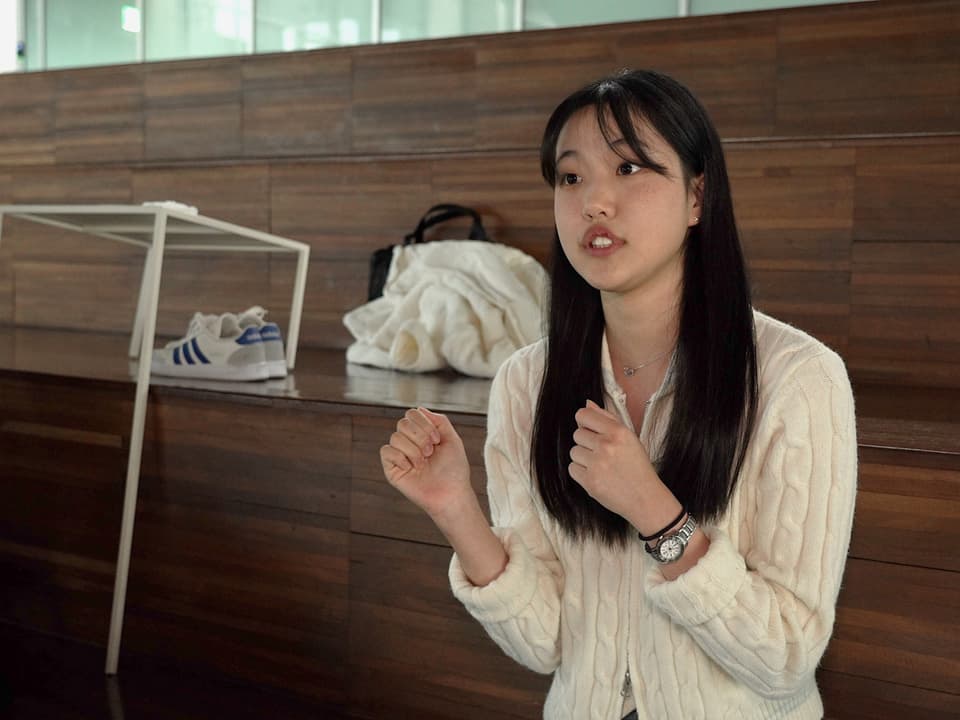 Junge Frau spricht gestikulierend in einem Raum mit Holzwand und abgelegten Gegenständen im Hintergrund.