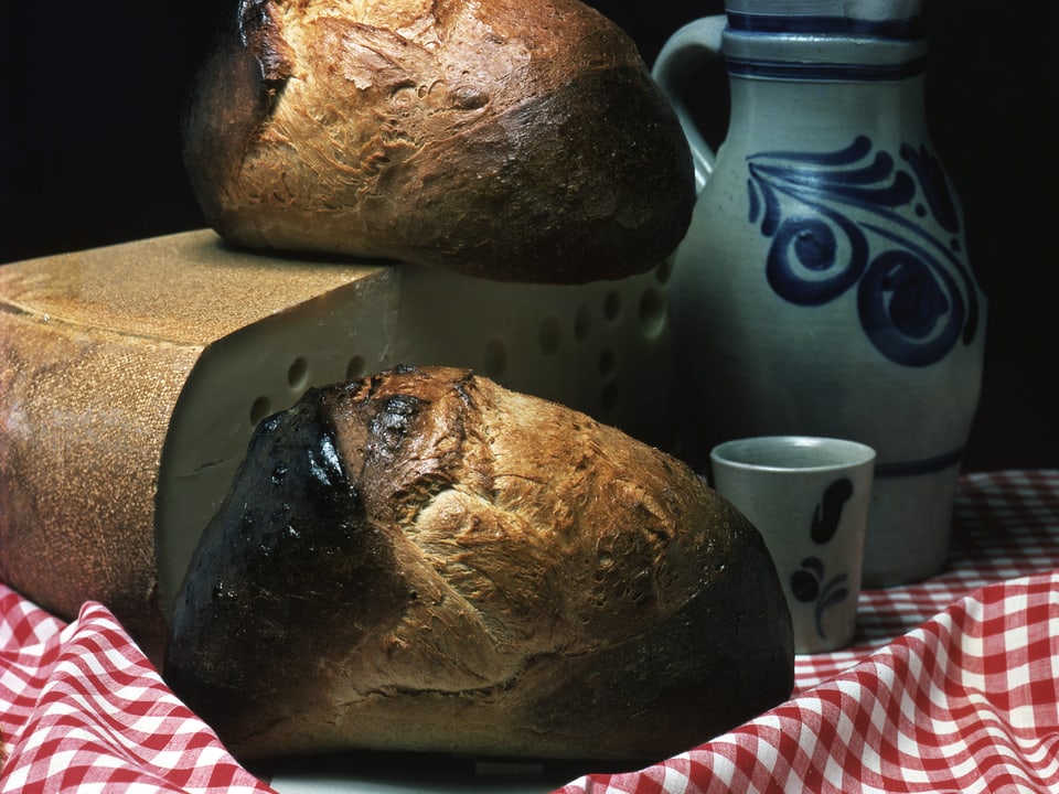 Zwei Laibe Brot auf einen karierten Tischtuch vor und auf einem Laib Käse.