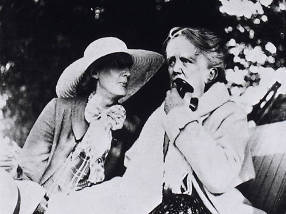 schwarzweiss Foto zwei älterer Frauen, links mit Hut und Schleife, rechts im Mantel, hält etwas in der Hand.