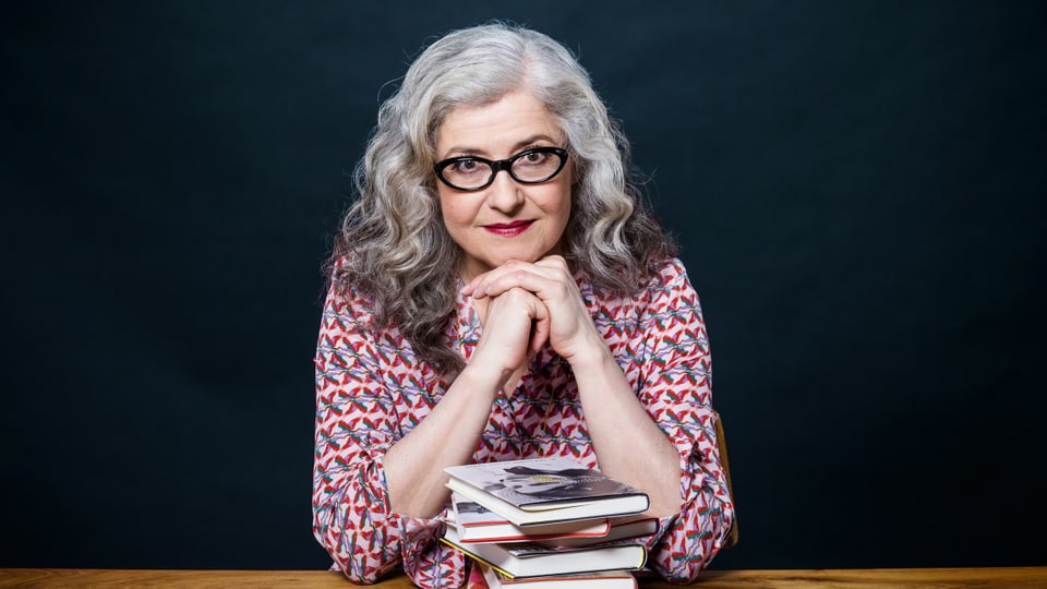 Eine ältere Frau mit grauen langen Haaren und schwarzer Brille mit Büchern auf dem Tisch lächelt freundlich.