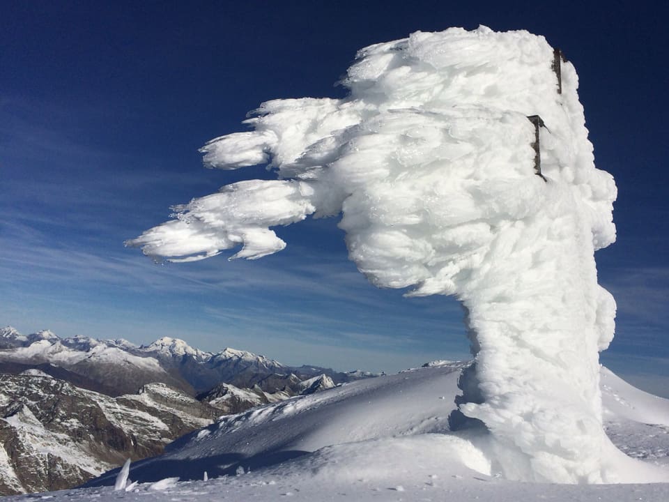 Stahlblauer Himmel, ein Gipfel mit Blick auf verschneites Bergpanorama. Das Gipfelkreuz ist in Eis verpackt. Das Eis zeigt noch die Spuren der Windrichtung. Es bildet eine Art Fahne. 