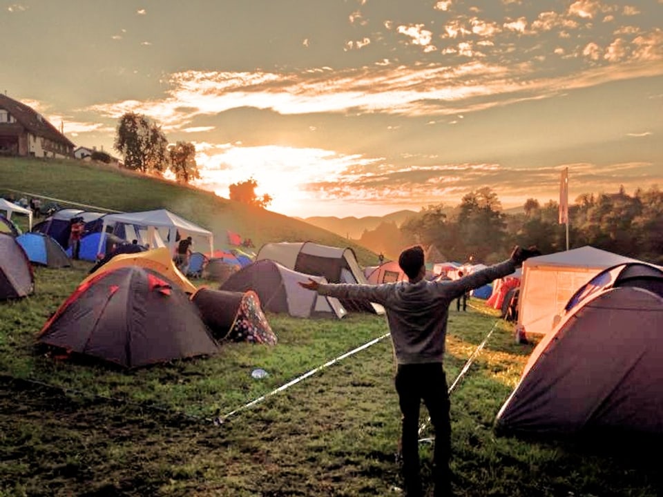 Zelte stehen am Hügel bei Sonnenaufgang, ein Mann streckt seine Arme der Sonne entgegen.