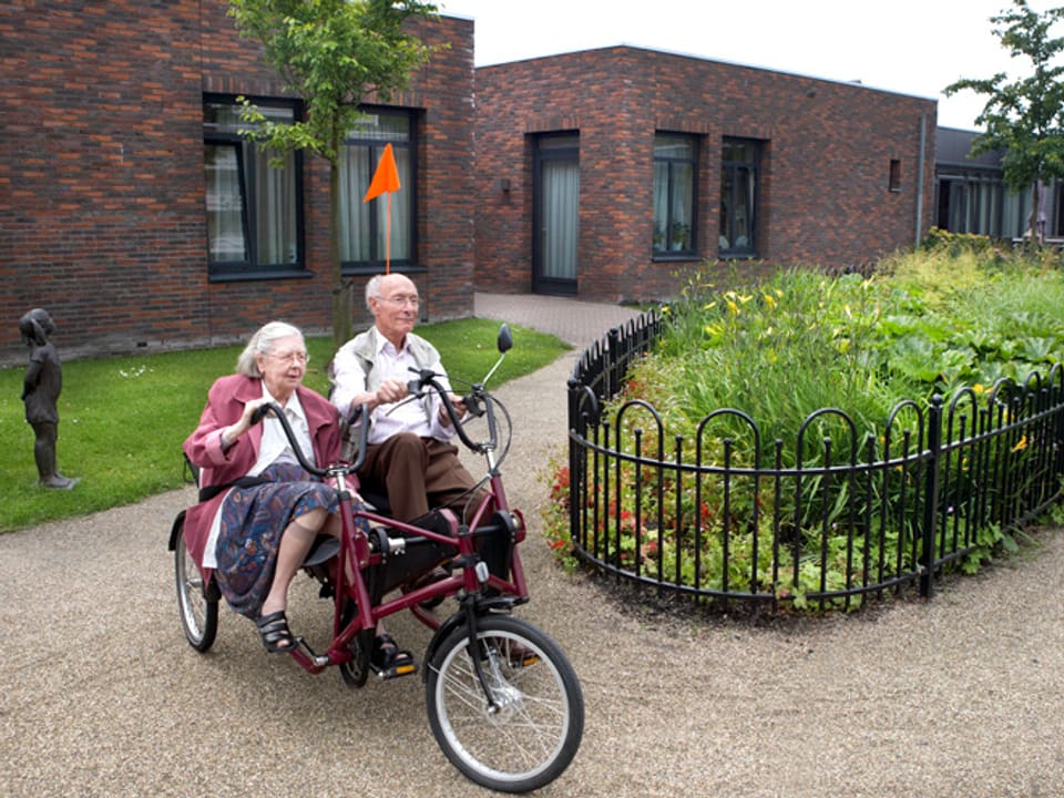 Ein älteres Paar fährt auf einem Velo mit Beifahrersitz durch ein Quartier.