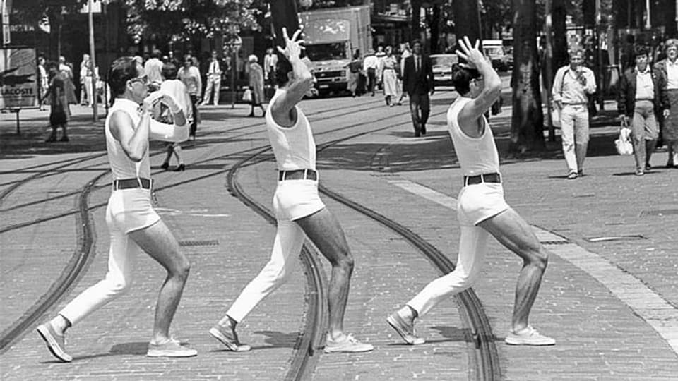 Drei Männer mit einbeinigen Hosen laufen auf Tramgleisen.